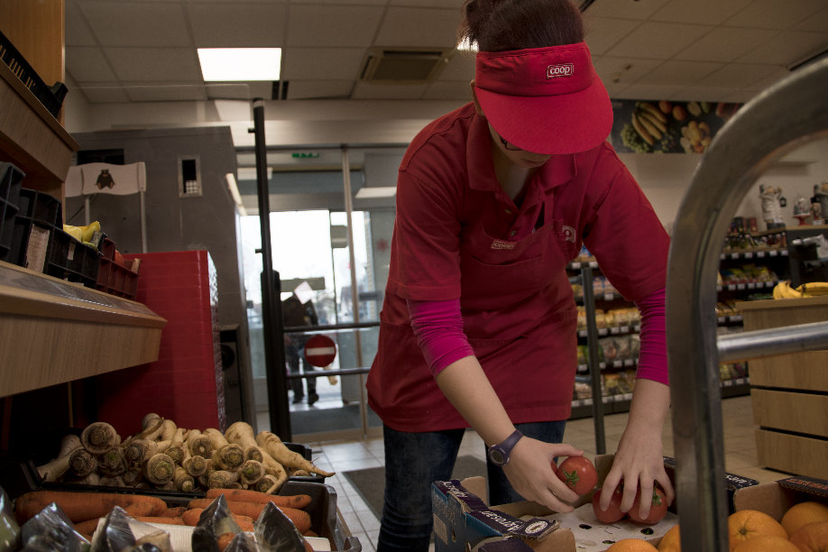 A képen egy hölgy látható munka közben, aki egy boltban éppen zöldségeket pakol.  